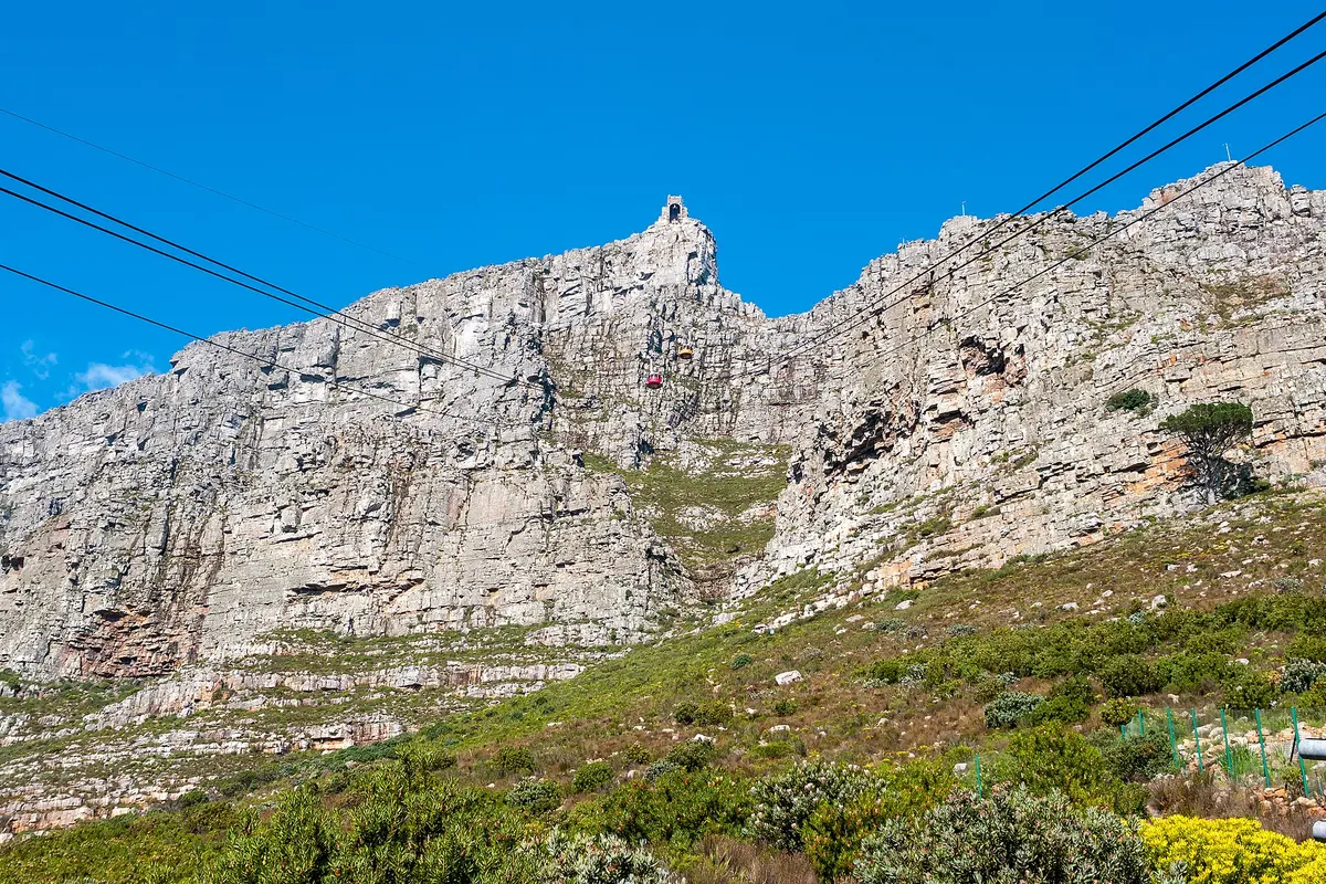 Two cable cars, one red and one yellow, on the cables below the upper station on the rocky skyline of Table Mountain under a blue sky