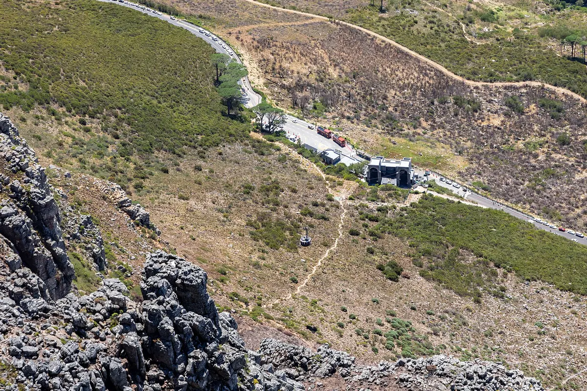 The lower cable station of Table Mountain seen from above, beside a road along the mountainside with parked cars and two red buses, and a cable car on the cables